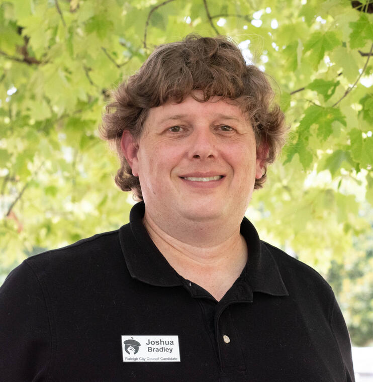 This is Josh's headshot. He is pictured smiling in front of a tree with a black polo shirt. He is wearing a metal nametag that says "Joshua Bradley Candidate for Raleigh City Council At-Large."